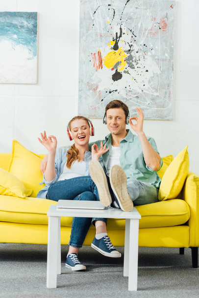 Wesoła para pokazuje ok gest słuchając muzyki w słuchawkach na kanapie w domu  - Zdjęcie, obraz