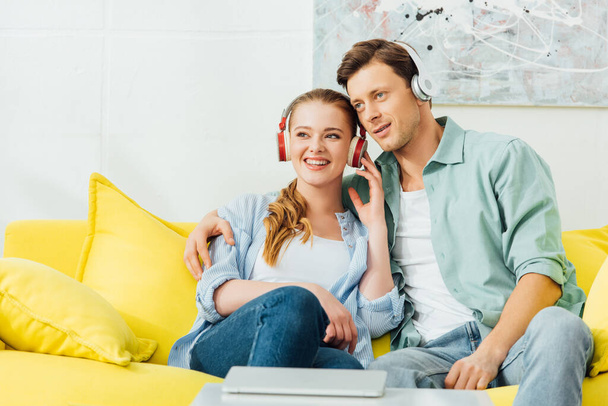 Selektiver Fokus des Mannes umarmt lächelnde Freundin mit Kopfhörern auf Couch neben Laptop auf Couchtisch  - Foto, Bild