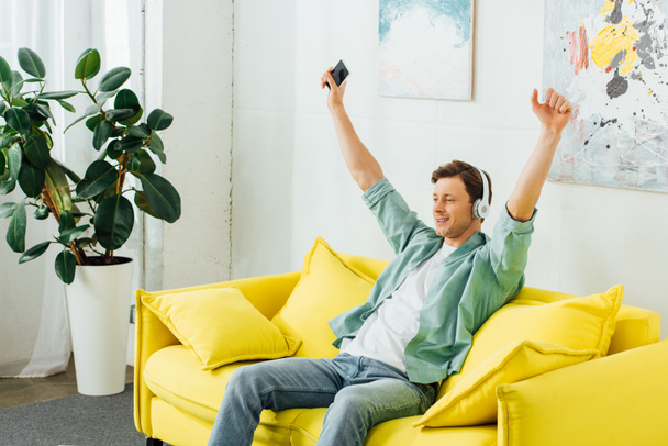 Excited man in headphones listening music and holding smartphone on couch at home  - Foto, Bild