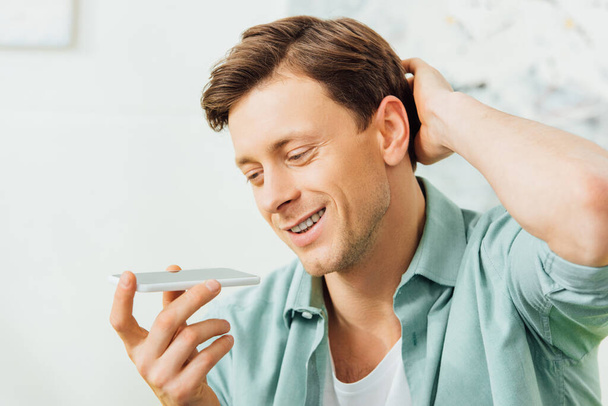 Handsome man smiling while using speakerphone on smartphone at home  - Fotó, kép