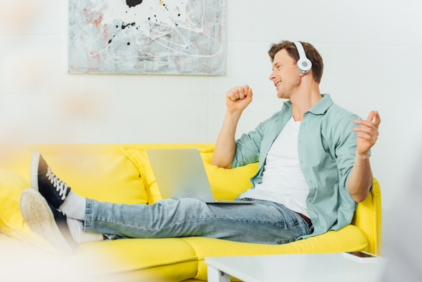 Side view of smiling man in headphones singing while sitting with laptop on sofa  - 写真・画像