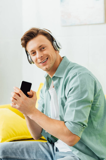 Side view of man in headphones smiling at camera and holding smartphone on couch  - Foto, Imagen