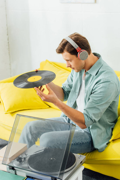 Side view of handsome man in headphones holding vinyl record near books and record player on coffee table  - Foto, afbeelding