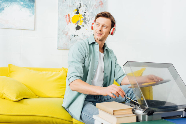 Smiling man in headphones looking away while using record player near books on coffee table - Photo, Image