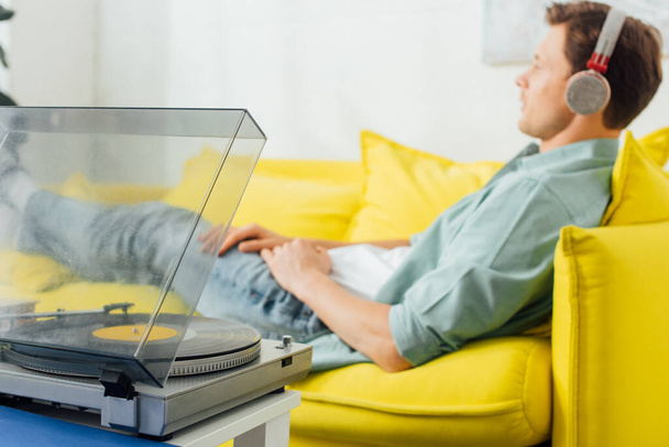 Selective focus of record player on coffee table and man in headphones lying on couch at home - Fotografie, Obrázek