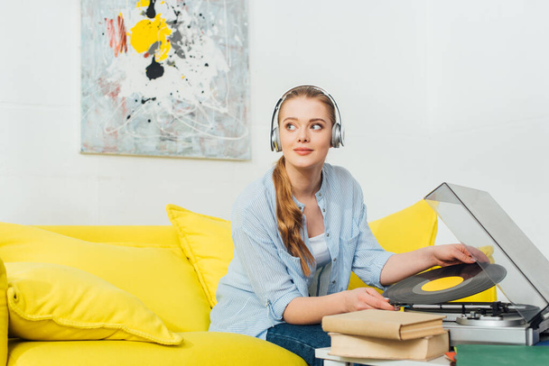 Beautiful woman in headphones holding vinyl record near books and  record player  on couch in living room - Foto, imagen