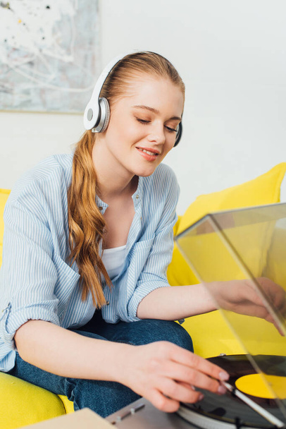 Selective focus of smiling woman in headphones using record player while sitting on sofa at home  - Fotó, kép