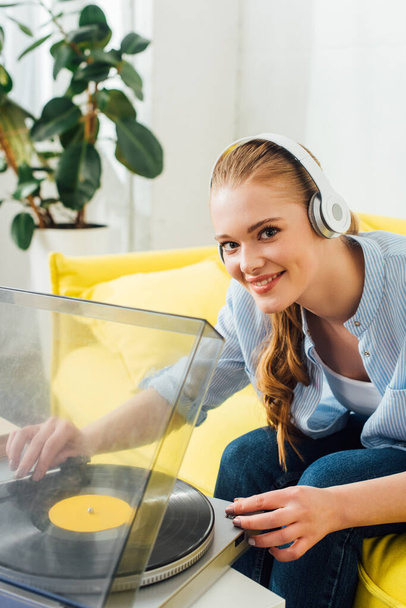 Smiling girl in headphones looking at camera near record player in living room - Photo, Image