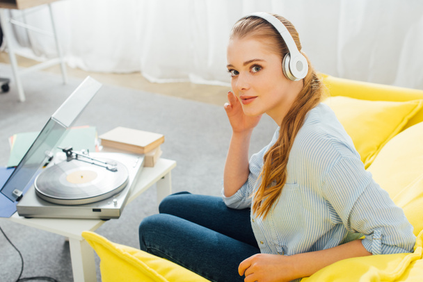 Zijaanzicht van de vrouw die naar de camera kijkt terwijl ze muziek luistert in een koptelefoon in de buurt van een platenspeler en boeken op salontafel - Foto, afbeelding