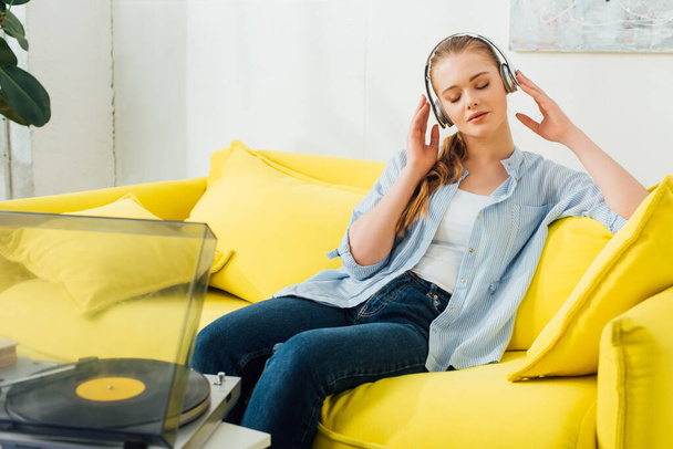 Beautiful woman listening music in headphones near record player in living room - Foto, afbeelding