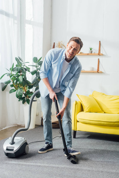 Handsome man smiling at camera while cleaning carpet with vacuum cleaner at home  - Fotó, kép