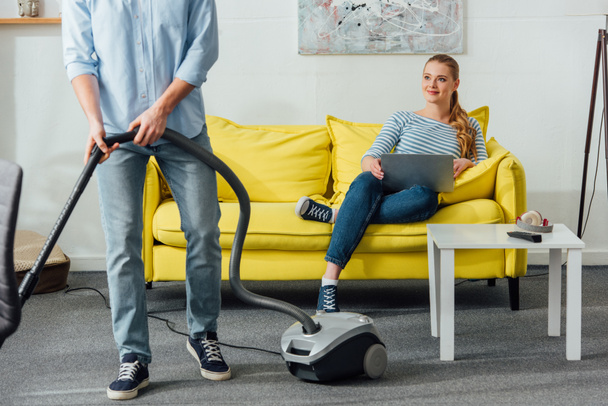 Smiling girl with laptop looking at boyfriend with vacuum cleaner in living room - Foto, Imagem