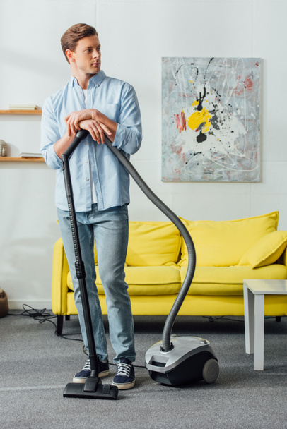 Handsome man standing near vacuum cleaner in living room  - Photo, Image