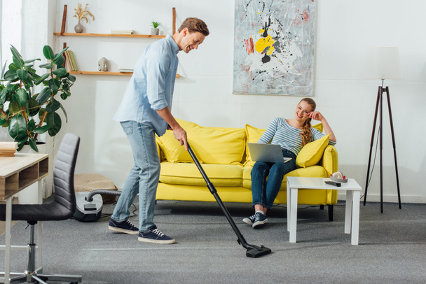 Smiling man cleaning carpet with vacuum cleaner near girlfriend using laptop on sofa at home  - Zdjęcie, obraz