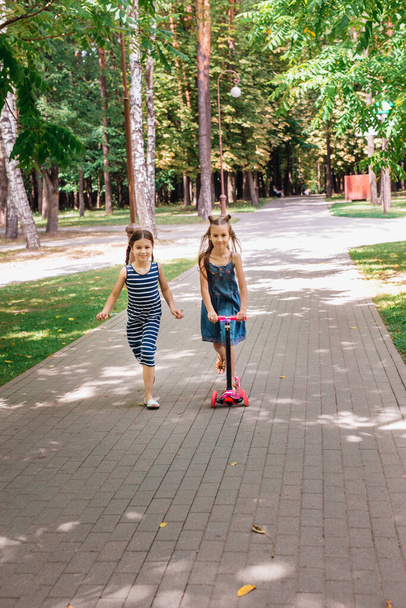 Two little girls riding scooters in summer park - Fotó, kép