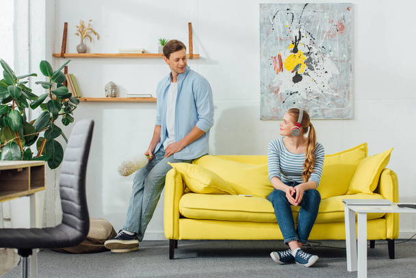 Muž s prachem štětec se usmívá na přítelkyni ve sluchátkách na gauči v obývacím pokoji - Fotografie, Obrázek
