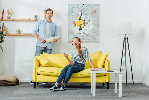 Woman in headphones with smartphone and boyfriend with dust brush smiling at camera in living room - Photo, image