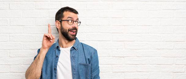 Hombre guapo con barba sobre la pared de ladrillo blanco con la intención de realizar la solución mientras levanta un dedo
 - Foto, imagen