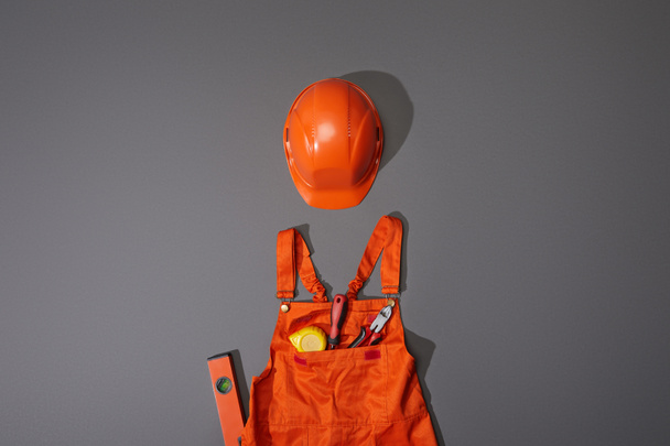 Draufsicht auf orangefarbenen Helm und Overall mit Maßband, Schraubenzieher und Zange auf grauem Hintergrund - Foto, Bild