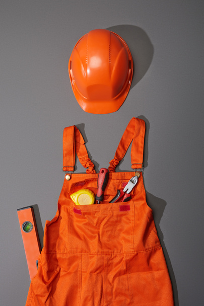 top view of orange helmet and overalls with measuring tape, screwdriver and pliers near spirit level on grey background - Foto, imagen