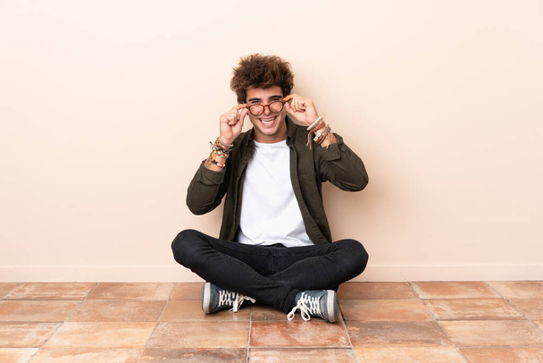 Jovem caucasiano sentado no chão com óculos e surpreso
 - Foto, Imagem