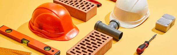 panoramic shot of bricks, helmets, spirit level, pipe connector, folding ruler, and screwdriver on yellow background - Photo, Image
