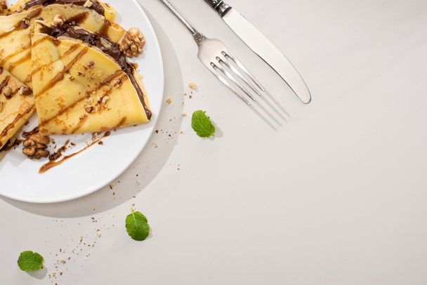 top view νόστιμες κρέπες με επικάλυψη σοκολάτας και καρύδια στο πιάτο κοντά σε μαχαιροπήρουνα και φύλλα δυόσμου σε γκρι φόντο - Φωτογραφία, εικόνα
