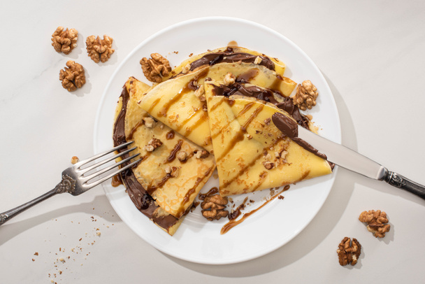 вид сверху на вкусные блины с шоколадом и грецкие орехи на тарелке со столовыми приборами на сером фоне
 - Фото, изображение