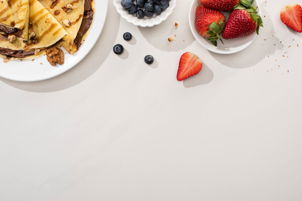 top view of tasty crepes with chocolate spread and walnuts on plate near bowls with blueberries and strawberries on grey background - Photo, Image