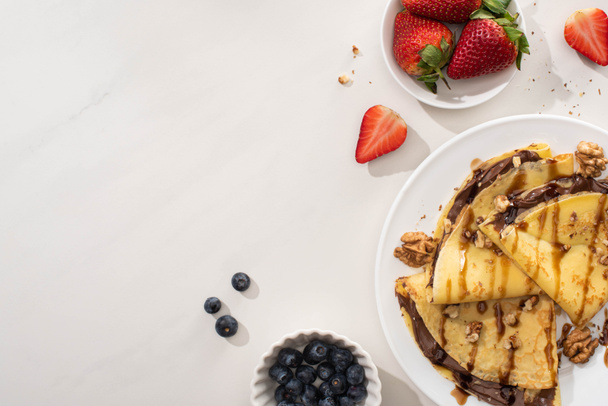 felülnézet ízletes palacsinta csokoládé terjedt és dió tányéron közelében tálak áfonya és eper szürke alapon - Fotó, kép