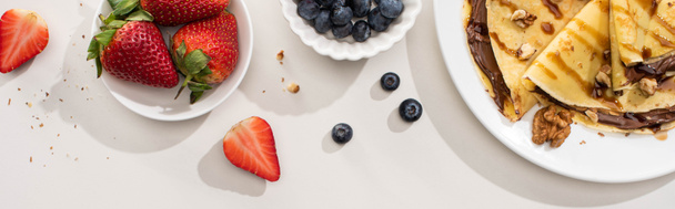 Draufsicht auf leckere Crêpes mit Schokoladenaufstrich und Walnüssen auf Teller in der Nähe von Schüsseln mit Blaubeeren und Erdbeeren auf grauem Hintergrund, Panoramaaufnahme - Foto, Bild