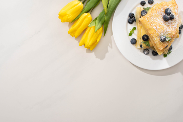 top view of tasty crepes with blueberries and mint on plate near yellow tulips on grey background - Photo, Image
