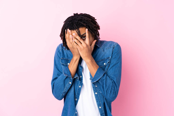 Young African American man with jean shirt over isolated pink background covering eyes and looking through fingers - Фото, изображение