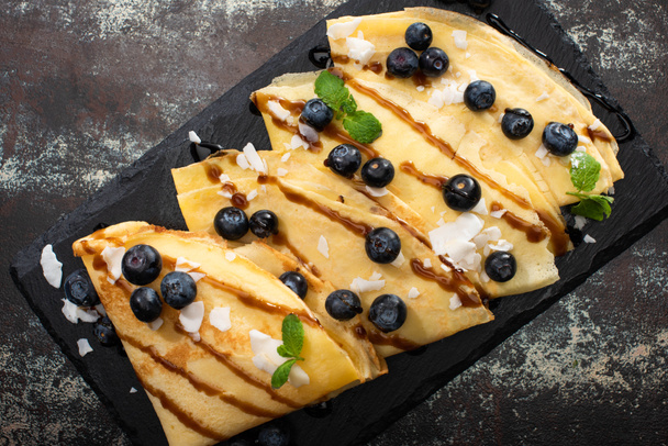 top view of tasty crepes with blueberries, mint and coconut flakes served on board on textured background - Fotoğraf, Görsel