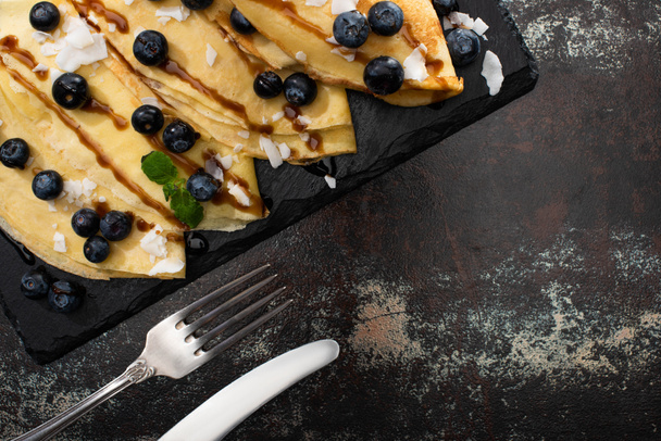 top view of tasty crepes with blueberries, mint and coconut flakes served on board near cutlery on textured background - Φωτογραφία, εικόνα