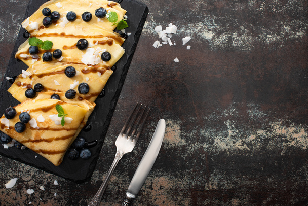 top view of tasty crepes with blueberries, mint and coconut flakes served on board near cutlery on textured background - Photo, Image