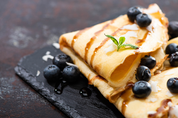 close up view of tasty crepes with blueberries, mint and coconut flakes served on board on textured background - Photo, Image
