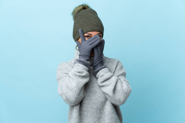 Молодая уругвайская девушка в зимней шляпе изолирована на синем фоне закрывая глаза и глядя сквозь пальцы
 - Фото, изображение