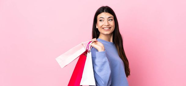 Mladá brunetka dívka přes izolované růžové pozadí drží nákupní tašky a ohlíží se - Fotografie, Obrázek