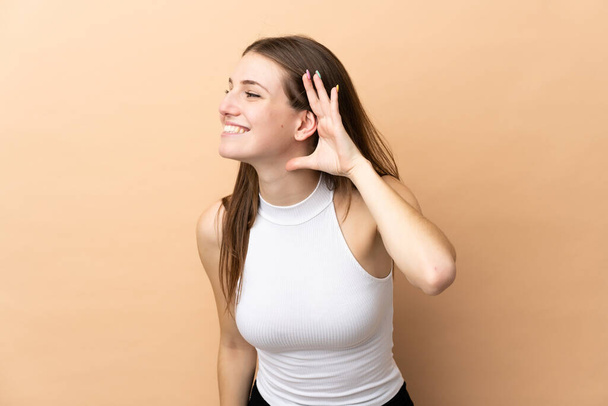 Νεαρή καυκάσια γυναίκα απομονωμένη σε μπεζ φόντο ακούγοντας κάτι βάζοντας το χέρι στο αυτί - Φωτογραφία, εικόνα