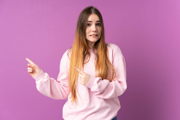 Young caucasian woman isolated on purple background frightened and pointing to the side - Photo, image