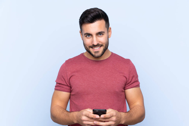 Joven hombre guapo con barba sobre fondo azul aislado enviando un mensaje con el móvil - Foto, imagen