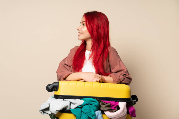 Traveler girl with a suitcase full of clothes isolated on beige background looking side - Photo, Image