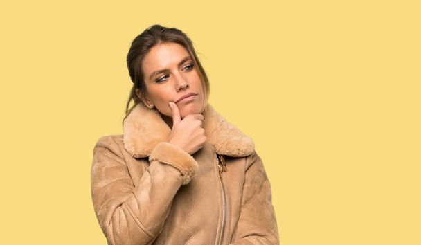 Blonde woman with a coat thinking over isolated yellow background - Photo, Image