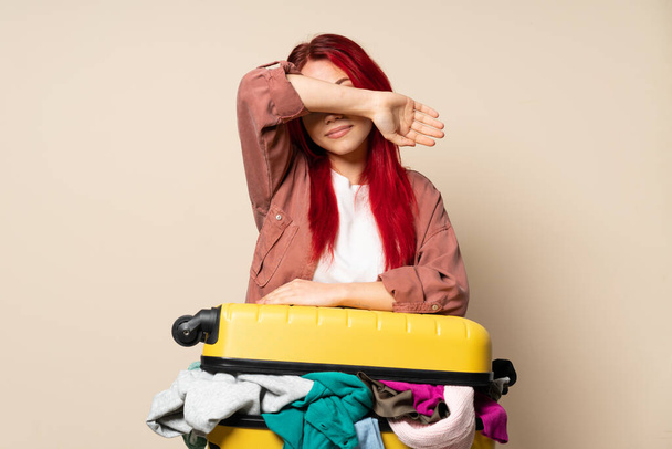 Chica viajera con una maleta llena de ropa aislada sobre fondo beige cubriendo los ojos con las manos
 - Foto, imagen