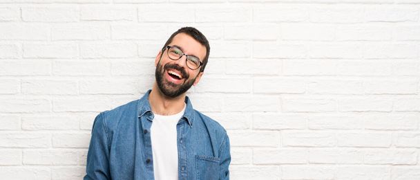 Όμορφος άντρας με γενειάδα πάνω από λευκό τούβλο τοίχο χαμογελώντας - Φωτογραφία, εικόνα