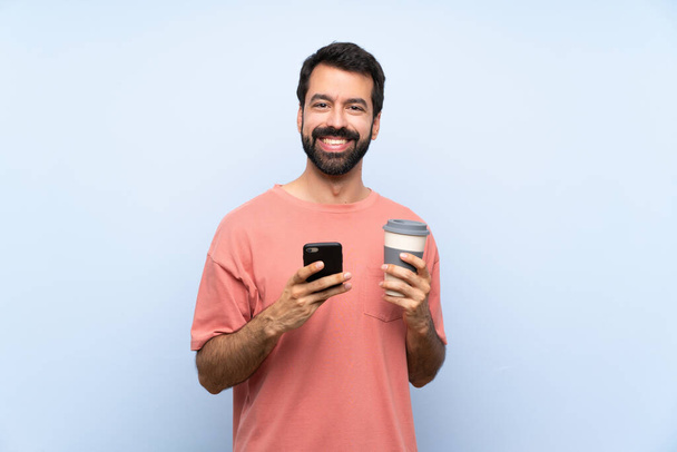 Young man with beard holding a take away coffee over isolated blue background sending a message with the mobile - Fotoğraf, Görsel