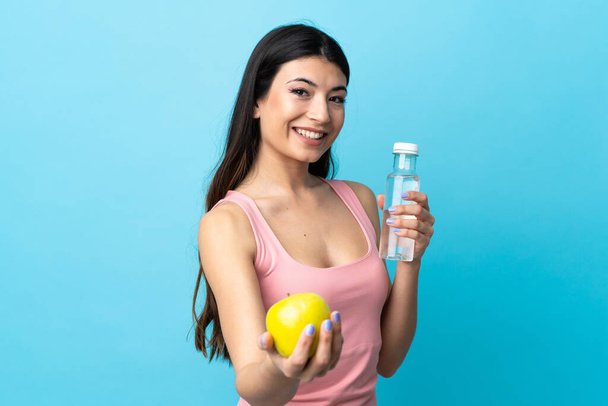 Mladá brunetka dívka přes izolované modré pozadí s jablkem a s lahví vody - Fotografie, Obrázek