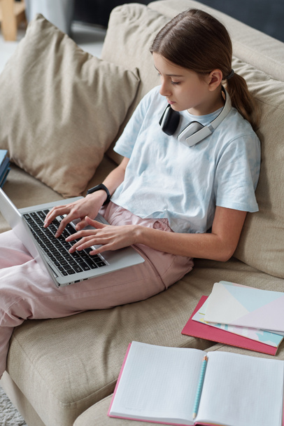 Bovenaanzicht van geconcentreerde tiener typen op laptop tijdens het werken aan school onderzoeksproject thuis - Foto, afbeelding