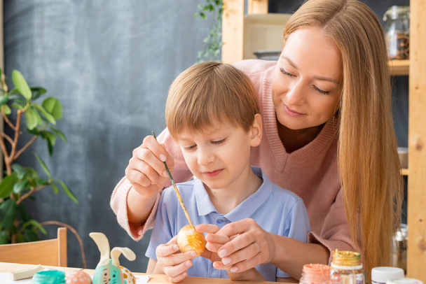 Attractive young mother using paintbrush while helping son to paint egg for Easter decorations - Photo, Image